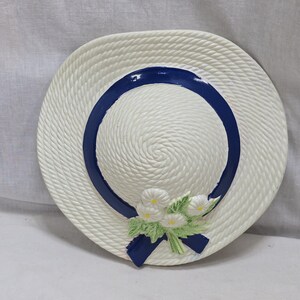 May include: White ceramic wall decor shaped like a straw hat with a blue ribbon and white flowers.