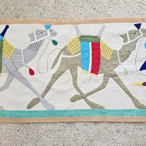 May include: A rectangular textile featuring three camels with colorful saddles and a figure in traditional attire. The camels are depicted in shades of gray and tan, with the figure running alongside them, holding a whip. The border is a light brown.
