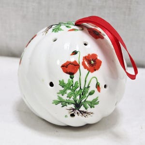 Puede incluir: Adorno de cerámica blanca con una cinta roja y un diseño de flores de amapola. El adorno tiene forma redonda con pequeños agujeros y una textura retorcida. Las amapolas son rojas con hojas verdes.