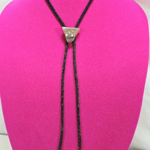 May include: A bolo tie with a triangular, light-colored stone pendant. The pendant is set on a black braided cord with silver-toned metal tips. The tie is displayed on a pink mannequin.