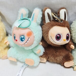 May include: Two plush toy figures, one in light green and the other in brown, each with a unique design. The light green figure has blue eyes and a bunny-like hood. The brown figure has large pink eyes and a furry texture. Both have keychains.
