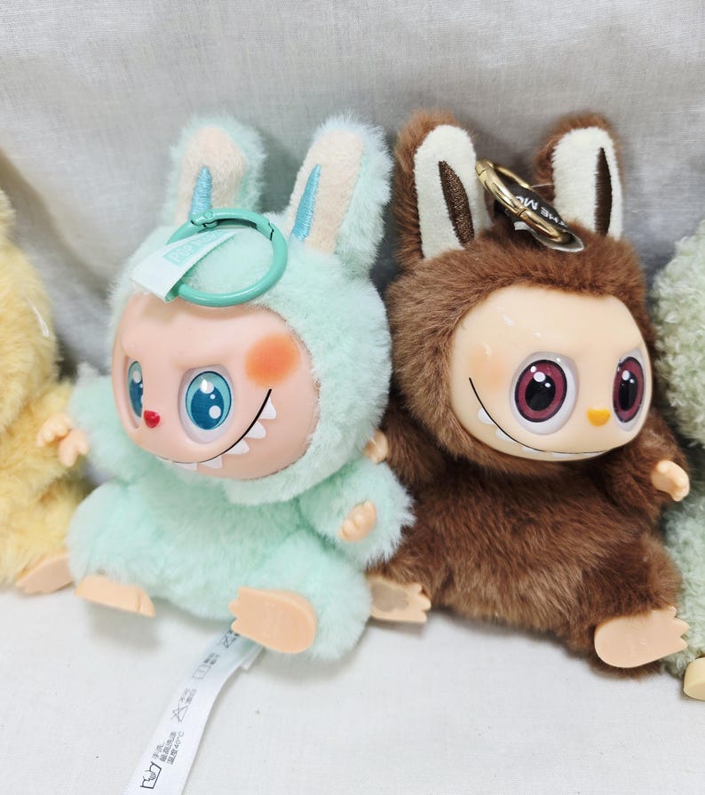 May include: Two plush toy figures, one in light green and the other in brown, each with a unique design. The light green figure has blue eyes and a bunny-like hood. The brown figure has large pink eyes and a furry texture. Both have keychains.