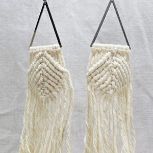 Puede incluir: Un par de pendientes de macramé color crema. Cada pendiente presenta una parte superior triangular plateada y un centro tejido en forma de diamante. Debajo del centro, cuelgan largas hebras deshilachadas del mismo material color crema.