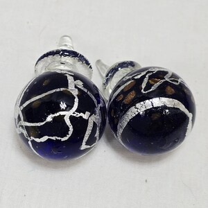 May include: Two decorative glass bottle stoppers. The stoppers have a dark blue, spherical body with silver and gold accents. The tops are clear glass with a silver band. The stoppers are on a white surface.
