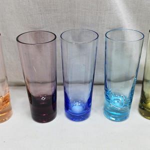 May include: Five tall, slender drinking glasses in a row, each with a different color. The colors are orange, purple, blue, teal, and olive green. The glasses are made of clear glass.