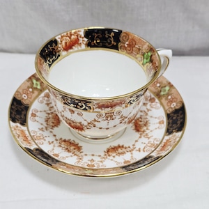 May include: A vintage teacup and saucer set. The white porcelain cup and saucer feature a floral pattern in shades of brown, orange, and black, with gold trim. The cup has a handle and sits on a matching saucer.