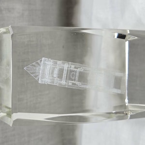 Könnte beinhalten: Ein klarer, rechteckiger Briefbeschwerer aus Glas mit einer dreidimensionalen, lasergravierten Abbildung eines Schiffes im Inneren. Das Glas hat abgeschrägte Kanten und eine glatte, polierte Oberfläche, die die detaillierten weißen Linien des Schiffes zeigt.