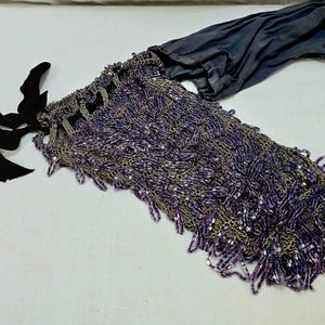 May include: A vintage evening bag with a beaded design in shades of purple and silver. The bag features a long, fringed body, a black ribbon tie, and a dark grey fabric lining.