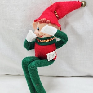 May include: A felt elf ornament featuring a red hat with a gold bell, a red torso, and green limbs. The elf has white gloves and white feet, a gold trim collar, and a painted face with blue eyes. A gold string is attached to the top of the hat.