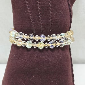 May include: Two-strand bracelet with iridescent, faceted beads. The beads are clear with hints of yellow and purple. The bracelet is wrapped around a dark burgundy fabric.