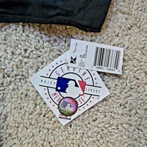 May include: A black garment with a tag attached. The tag features the Major League Baseball logo, a holographic sticker, and a barcode. The tag also includes text such as "GENUINE" and "MAJOR LEAGUE".