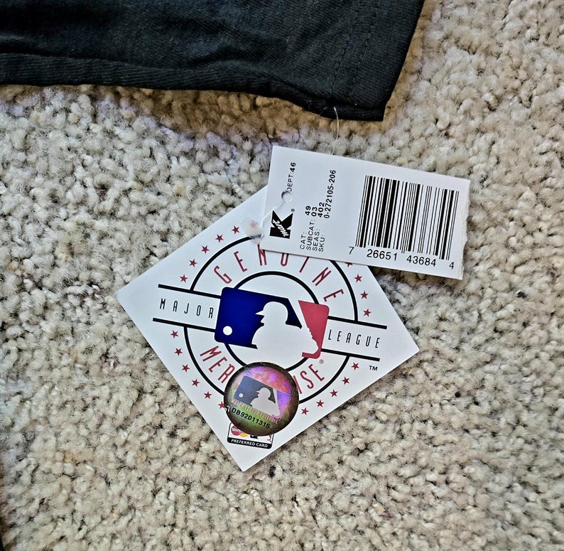 May include: A black garment with a tag attached. The tag features the Major League Baseball logo, a holographic sticker, and a barcode. The tag also includes text such as "GENUINE" and "MAJOR LEAGUE".