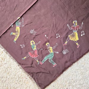 Puede incluir: Una tela marrón con un diseño de cuatro personas bailando. Las personas están delineadas en negro y tienen acentos amarillos, verdes y rojos. La tela tiene un borde de flecos.