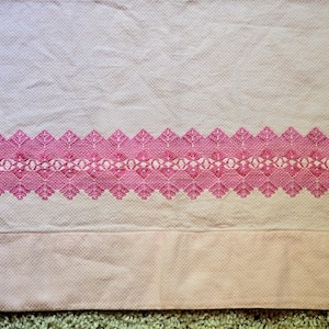May include: A light pink textile with a detailed, embroidered border in a darker shade of pink. The border features a repeating geometric and floral pattern. The fabric has a textured appearance.