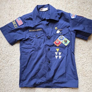 May include: A navy blue Boy Scouts of America uniform shirt with short sleeves. The shirt features patches with the American flag, the words "DEN 3", and various merit badges. The shirt has two chest pockets and black buttons.