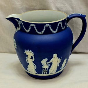 May include: A cobalt blue ceramic pitcher with a white interior and handle. The pitcher features white bas-relief figures and floral designs. The rim is decorated with a white garland design.