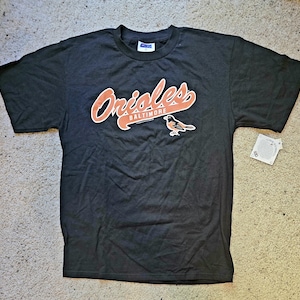 May include: Black t-shirt featuring the Baltimore Orioles logo in orange and white. The logo includes the word "Orioles" in a script font, the city name "Baltimore", and a bird graphic. A tag is attached to the shirt.