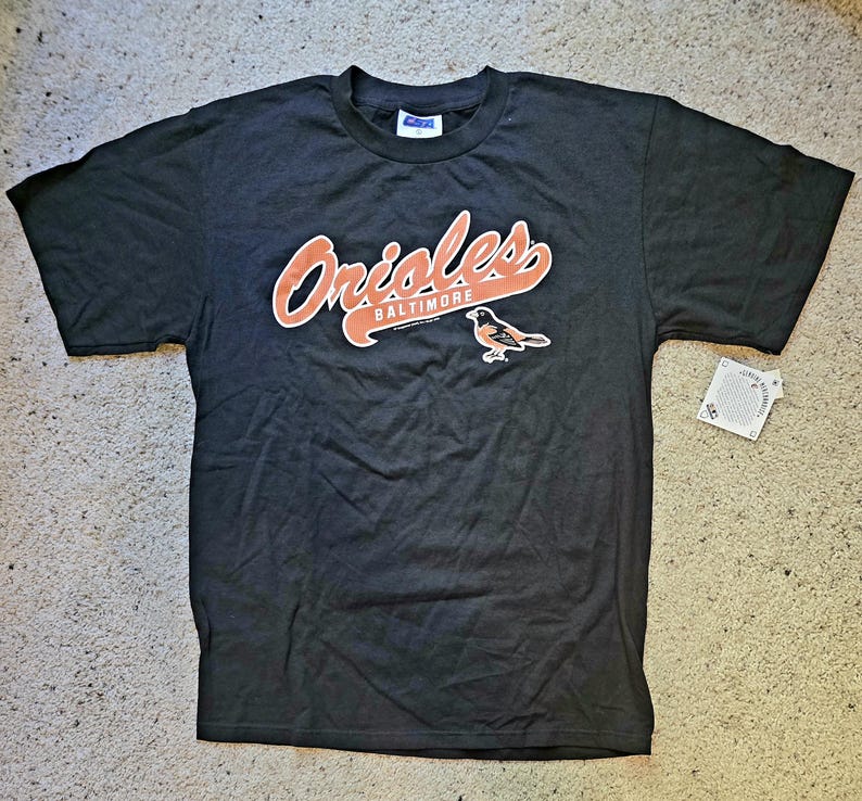 May include: Black t-shirt featuring the Baltimore Orioles logo in orange and white. The logo includes the word "Orioles" in a script font, the city name "Baltimore", and a bird graphic. A tag is attached to the shirt.