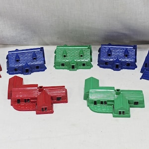 May include: A collection of ten small plastic game pieces shaped like buildings, including churches and houses in red, blue, and green. The set features various architectural designs with molded details such as roofs, windows, and doors, arranged in a group.