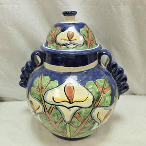 Könnte beinhalten: Eine blaue und weiße Keramikvase mit Deckel, verziert mit weißen Blumen und grünen Blättern. Die Vase hat zwei Griffe an den Seiten.