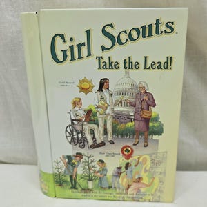 Puede incluir: Una caja de metal verde y blanca con un diseño de Girl Scouts Take the Lead!. La caja presenta ilustraciones de Girl Scouts en diversas actividades, incluyendo plantar árboles, participar en una carrera en silla de ruedas y reunirse con una política. La caja también incluye el texto "Gold Award 1960-Present" y "First Class Award 1960-1980".