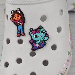 May include: Two colourful shoe charms attached to a white foam clog. One charm depicts a character with brown hair and cat ears wearing a pink jacket and blue jeans. The other charm features a teal cat with a patterned tail and star accents.