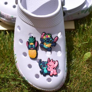 May include: White clogs with floral print detailing, decorated with colourful cartoon character charms. The charms feature a pineapple and Stitch characters in blue, pink, and yellow. The clogs are on grass.