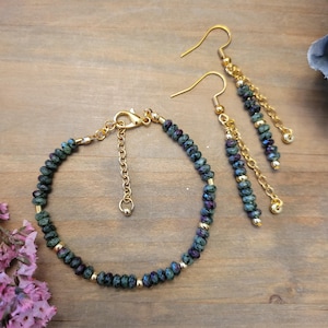 May include: A jewelry set featuring a bracelet and earrings. The bracelet and earrings have dark green and purple beads with gold accents. The earrings hang from gold hooks with dangling chains. The jewelry is displayed on a wooden surface.