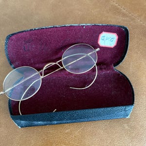 May include: A pair of vintage gold-tone metal spectacles with oval lenses and curved temple arms, resting inside an open black hard-shell case lined with deep burgundy velvet. A small rectangular white sticker with blue handwritten text is attached.