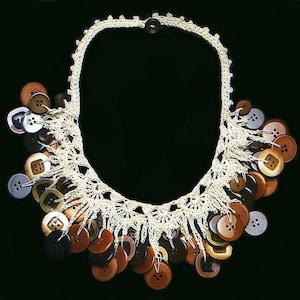 May include: A cream-coloured crochet necklace featuring a variety of brown, silver, and tan buttons. The buttons are attached to the chain with thread, creating a unique, textured design. The necklace is displayed against a black background.