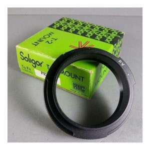 Puede incluir: Un anillo adaptador de montura Soligor T-2 negro con el texto "PV" en el lateral. El adaptador está delante de una caja verde con el logotipo de Soligor y el texto "T-2 MOUNT". La caja también tiene el texto "MADE IN JAPAN".