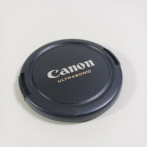 Peut inclure: Un bouchon d'objectif Canon noir avec le nom de la marque et "ULTRASONIC" en lettres argentées. Le bouchon rond a une surface légèrement texturée et trois petits clips sur le bord.
