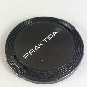 Peut inclure: Un bouchon d'objectif d'appareil photo noir avec le mot "PRAKTICA" imprimé en blanc. Le bouchon est rond avec un bord strié et une petite section qui semble être une charnière ou un mécanisme d'ouverture. Il est pour un objectif d'appareil photo.