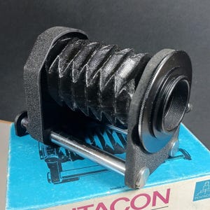 May include: Black bellows camera extension tube with a silver metal rod and a black plastic base. The tube is sitting on a blue box with the word "PENTACON" printed on it.