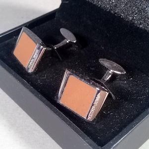 May include: A pair of square cufflinks featuring a polished silver-tone metal frame and a smooth tan-colored inlay. Each cufflink is engraved with the text HUGO BOSS along the side and is presented in a black box with a black velvet interior lining.