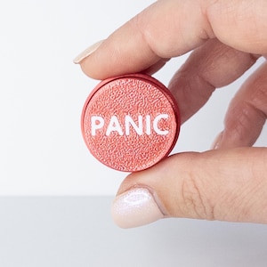 Puede incluir: Un pequeño botón redondo rojo con la palabra "PANIC" en blanco. El botón se sostiene entre los dedos y tiene una superficie texturizada. El fondo es blanco liso.
