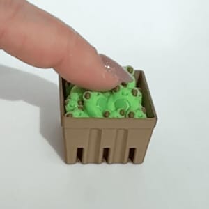 以下が含まれることがあります： 茶色のミニチュアバスケットには、茶色の斑点のある小さな明るい緑色のカエルの置物が入っています。指がそっとカエルに触れています。バスケットは長方形で、側面に垂直のスロットがあります。