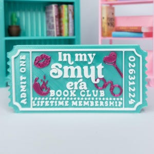 May include: A teal and white ticket with the text "In my Smut era BOOK CLUB LIFETIME MEMBERSHIP". The ticket features a pink lip print, a heart-shaped key, and handcuffs.