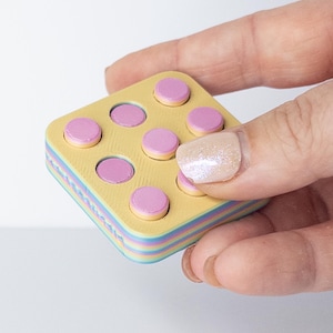 May include: A small, square, wooden toy with a yellow top and a rainbow-colored side. The top has eight pink and yellow circular pieces. The toy is held in a hand.