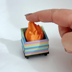May include: A 3D-printed miniature orange flame sits inside a colorful, striped box. The box is a mix of pastel colors, including blue, green, yellow, and pink. The flame is being held by a finger.