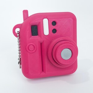 May include: A bright pink, 3D-printed camera keychain. The camera has a square body with a lens, flash, and viewfinder details. A silver ball chain is attached for hanging. The camera is a solid, vibrant pink color.