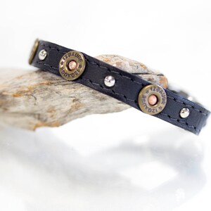 May include: Black leather bracelet with silver studs and brass shotgun shell casings. The casings have the text "12 Gauge" engraved on them.