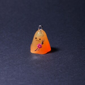 Può includere: Un piccolo ciondolo in resina arancione con un anello color argento. Il ciondolo presenta un volto da cartone animato e un motivo floreale rosa. Un accessorio unico.