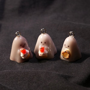 Może przedstawiać: Trzy małe, jasnofioletowe zawieszki w kształcie ducha ze srebrnymi pętelkami. Każdy duch trzyma miniaturową słodkość, w tym babeczkę i pączka. Te zawieszki są idealne do projektów biżuterii lub rękodzieła o tematyce Halloween.