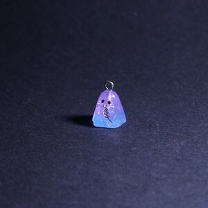 May include: A small, ghost-shaped pendant with a silver loop for hanging. The pendant is a gradient of blue and purple, with a small sprig and two red dots. The pendant is likely made of resin.