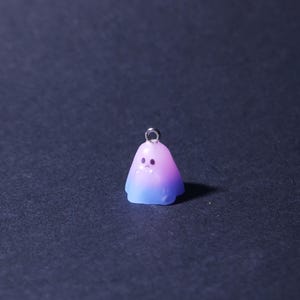 May include: A small, ghost-shaped charm with a gradient of blue and purple hues. The charm has a silver-colored loop at the top for hanging. The ghost has two small dot eyes and a small mouth.