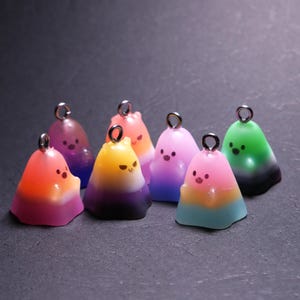May include: A collection of seven small, translucent ghost-shaped charms, each featuring a metal jump ring at the top. The charms display various gradient color combinations, including pink, orange, yellow, purple, blue, and green, with simple painted facial features.