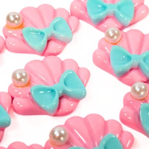 May include: Pink seashell-shaped embellishments with a light blue bow and a pearl accent. These decorative elements are ideal for crafting projects, with a glossy finish and a charming, whimsical design.