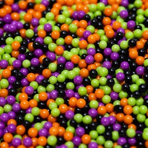 May include: A close-up shot of a pile of small, round sprinkles in various colors. The sprinkles are in shades of black, orange, purple, and green. These colorful sprinkles are likely for decorating baked goods.