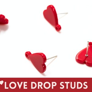 May include: Four pairs of red heart-shaped stud earrings with silver posts. The earrings are scattered on a white surface.  LOVE DROP STUDS is written in white text on a red background.
